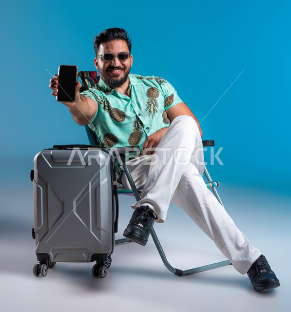 قضاء عطلة صيفية ترفيهيه والقيام بالرحلات الممتعه, استخدام الأجهزة والتقنيات الحديثة, عرض خدمات وتطبيقات السياحة والسفر, بورتريه لشاب عربي خليجي سعودي مبتسم يرتدي الزي الكاجوال والنظارة يجلس بجانب الحقيبة ويمسك الهاتف المحمول بشاشة سوداء فارغة, خلفية زرقاء