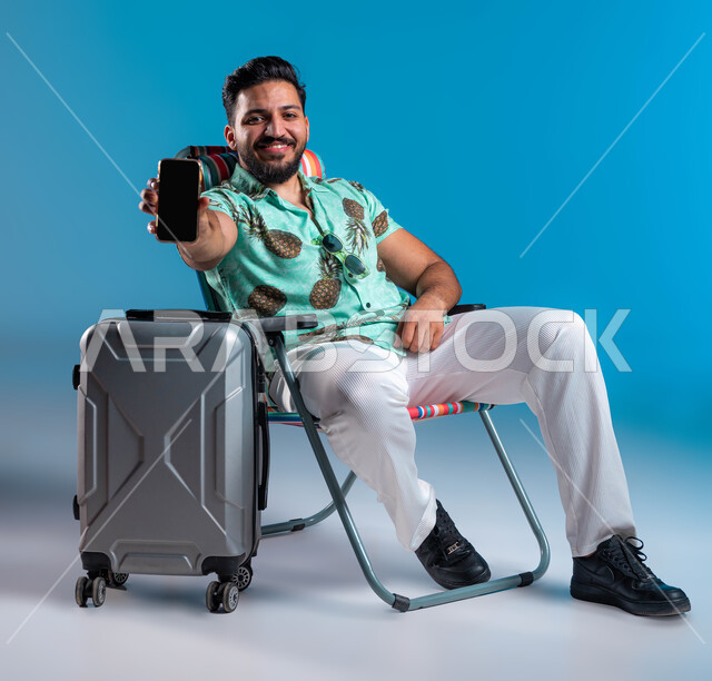 استخدام الأجهزة والتقنيات الحديثة والمتطورة, عرض خدمات وتطبيقات السياحة والسفر, رحلة شبابية ممتعه وقضاء عطلة صيفية ترفيهيه, بورتريه لشاب عربي خليجي سعودي مبتسم يرتدي الزي الكاجوال يجلس بجانب الحقيبة ويمسك الهاتف المحمول بشاشة سوداء فارغة, خلفية زرقاء