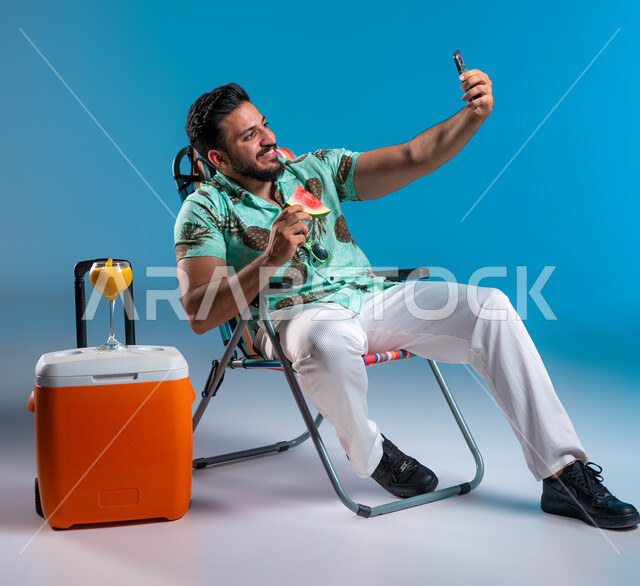 القيام بالبث المباشر عبر الهاتف المحمول, أهمية السفر والسياحة وقضاء أيام العطلة بالأماكن الساحلية السعودية, تناول الفواكه الصيفية المنعشة, بورتريه لشاب عربي خليجي سعودي مبتسم يرتدي الزي الكاجوال يقوم بالتقاط صور سيلفي تذكارية عبر الجوال, خلفية زرقاء