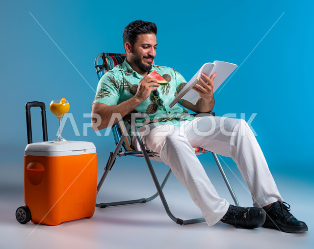 قضاء أيام العطلة في الرحلات السياحية, تناول الفواكه الصيفية المنعشة, ممارسة الهواية المفضلة في أوقات الراحة, بورتريه لرجل عربي خليجي سعودي مبتسم يرتدي الزي الكاجوال يقوم بقراءة كتاب ما, الاستمتاع بالعصائر الطبيعيه الباردة, خلفية زرقاء