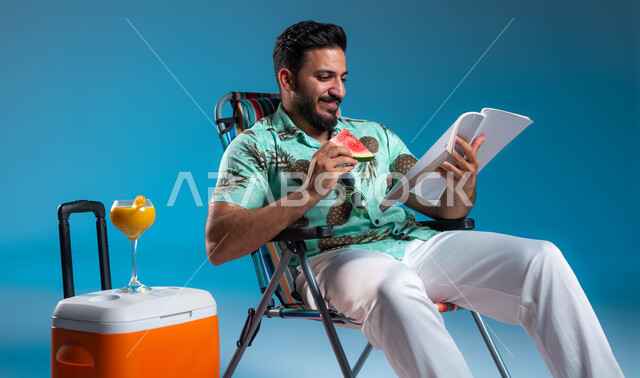 قضاء أيام العطلة في الرحلات السياحية, تناول الفواكه الصيفية المنعشة, ممارسة الهواية المفضلة في أوقات الراحة, بورتريه لرجل عربي خليجي سعودي مبتسم يرتدي الزي الكاجوال يقوم بقراءة كتاب ما, الاستمتاع بالعصائر الطبيعيه الباردة, خلفية زرقاء