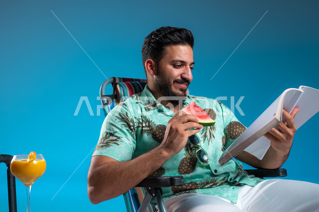 تناول الفواكه الصيفية المنعشة, ممارسة الهواية المفضلة في أوقات الراحة, بورتريه مقرب لرجل عربي خليجي سعودي مبتسم يرتدي الزي الكاجوال يقوم بقراءة كتاب ما, الاستمتاع بالمشروبات الباردة وقضاء أيام العطلة في الرحلات السياحية, خلفية زرقاء