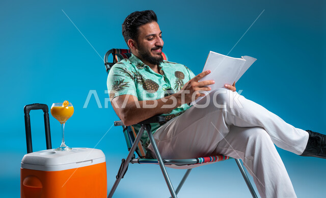 رحلة شبابية ممتعه وقضاء أوقات العطلة الصيفية بالسياحة الترفيهية, الجلوس براحة وممارسة الهوايات المفيدة في أوقات الفراغ, بورتريه لشاب عربي خليجي سعودي مبتسم يرتدي الزي الكاجوال يجلس على الكرسي ويقوم بقراءة كتاب ما, الاستمتاع بالمشروبات الباردة, خلفية زرقاء