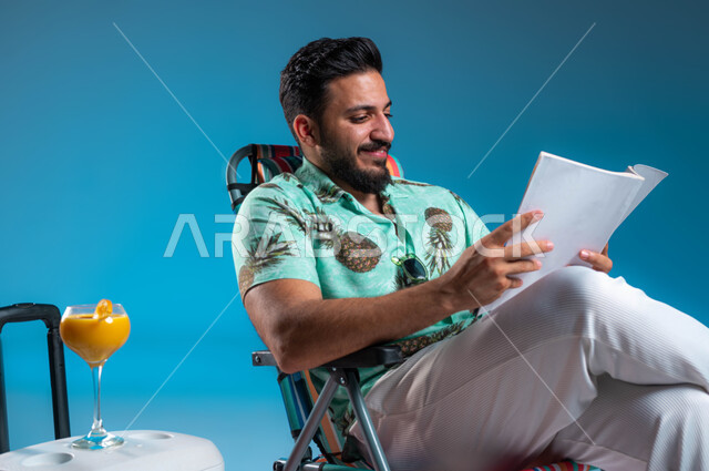 الجلوس براحة وممارسة الهوايات المفيدة في أوقات الفراغ, بورتريه لشاب عربي خليجي سعودي مبتسم يرتدي الزي الكاجوال يجلس على الكرسي ويقوم بقراءة كتاب ما, الاستمتاع بالمشروبات الباردة, رحلة شبابية ممتعه وقضاء أوقات العطلة الصيفية بالسياحة الترفيهية, خلفية زرقاء