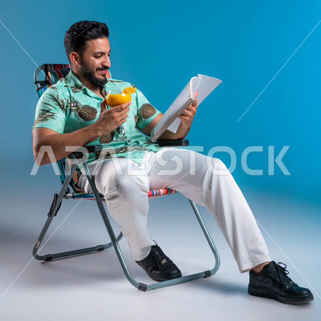 التعلم والثقافة وممارسة الهوايات المفيدة في أوقات الفراغ, بورتريه لشاب عربي خليجي سعودي مبتسم يرتدي الزي الكاجوال يجلس على الكرسي ويقوم بقراءة كتاب ما, الاستمتاع بالمشروبات الباردة, رحلة شابية ممتعه وقضاء أوقات العطلة الصيفية بالسياحة الترفيهية, خلفية زرقاء