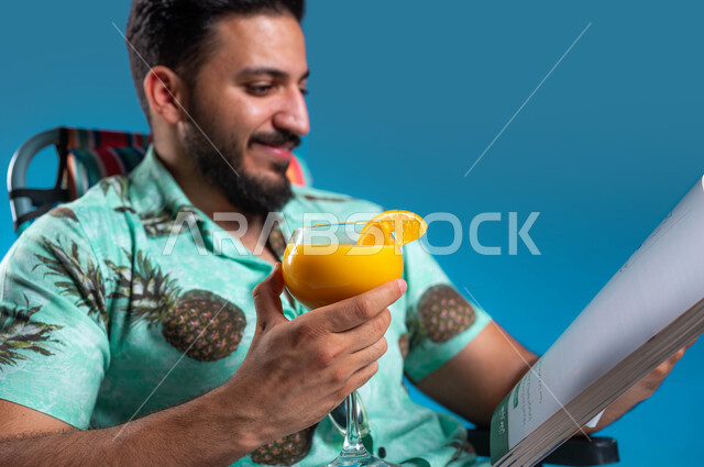 ممارسة الهوايات المفيدة في أوقات الفراغ, بورتريه مقرب لشاب عربي خليجي سعودي مبتسم يرتدي الزي الكاجوال يقوم بقراءة كتاب ما, الاستمتاع بالمشروبات الباردة, رحلة شابية ممتعه وقضاء أوقات العطلة الصيفية بالسياحة الترفيهية, خلفية زرقاء