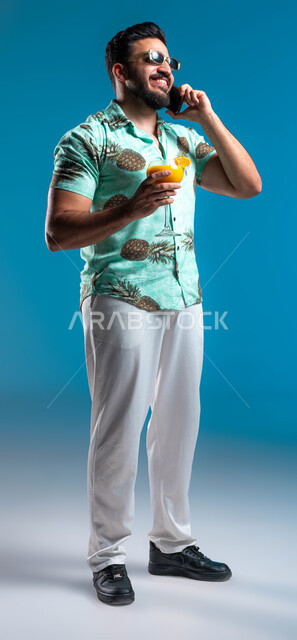 إجراء مكالمة هاتفية والتواصل مع الأصدقاء عبر الهاتف المحمول, قضاء أوقات العطلة الصيفية بالرحلات الشبابية الممتعه, بورتريه لشاب عربي خليجي سعودي مبتسم يرتدي الزي الكاجوال يستمتع بشرب العصير الطبيعي البارد بإيماءات الفرح, صورة الجسم بالطول الكامل, خلفية زرقاء