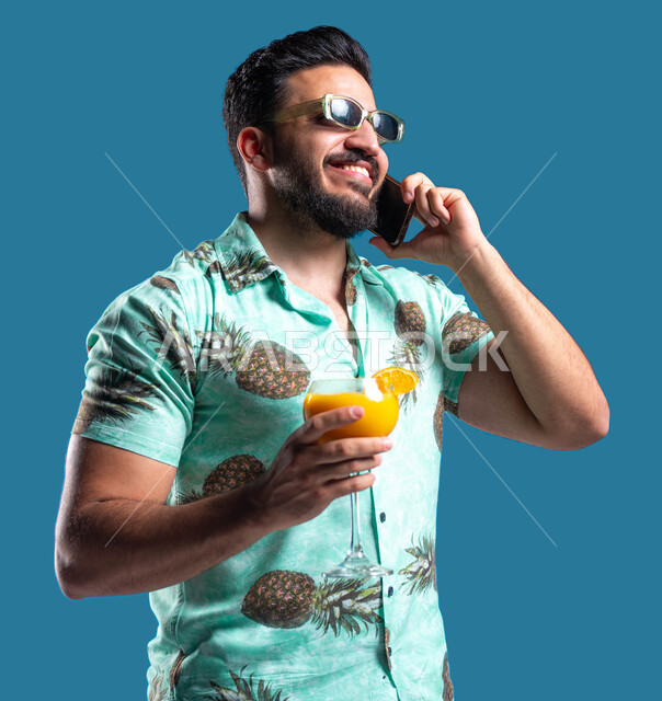 قضاء أوقات العطلة الصيفية بالرحلات الشبابية الممتعه, التواصل مع الأصدقاء عبر الهاتف المحمول, بورتريه مقرب لشاب عربي خليجي سعودي مبتسم يرتدي الزي الكاجوال يقوم بإجراء مكالمة هاتفيه عبر الجوال بإيماءات السرور, الاستمتاع بشرب العصير الطبيعي البارد, خلفية زرقاء