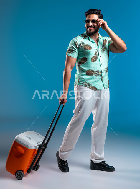 الاستمتاع بالمشروبات الباردة وقضاء وقت ممتع في العطلة الصيفية, السياحة في إحدى منتجعات السعودية, بورتريه لشاب عربي خليجي سعودي مبتسم يرتدي الزي الكاجوال والنظارة الشمسية يحمل حافظة العصائر وينظر إلى شيء ما, صورة الجسم بالكامل, خلفية زرقاء