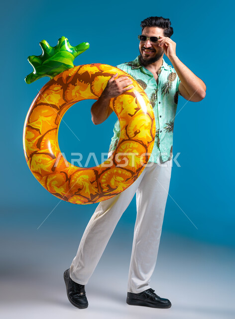 الوقوف والنظر إلى شيء ما, قضاء أوقات ممتعه في شواطئ السعودية بإيماءات السعادة والسرور, التسلية في إحدى المنتجعات الترفيهيه, بورتريه لشاب عربي خليجي سعودي مبتسم يرتدي الزي الكاجوال والنظارة يمسك بيده عوامة السباحة, صورة الجسم بالطول الكامل, خلفية زرقاء