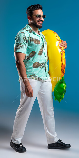 قضاء أوقات ممتعه والاستمتاع في شواطئ السعودية بإيماءات البشاشة والسرور, التسلية في إحدى المنتجعات الترفيهيه, بورتريه لشاب عربي خليجي سعودي مبتسم يرتدي الزي الكاجوال والنظارة يمسك بيده عوامة السباحة, صورة الجسم بالطول الكامل, خلفية زرقاء