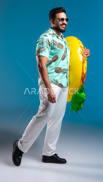 التسلية في إحدى المنتجعات الترفيهيه, بورتريه لشاب عربي خليجي سعودي مبتسم يرتدي الزي الكاجوال والنظارة يمسك بيده عوامة السباحة, قضاء أوقات ممتعه والاستمتاع في شواطئ السعودية بإيماءات البشاشة والسرور, صورة الجسم بالطول الكامل, خلفية زرقاء