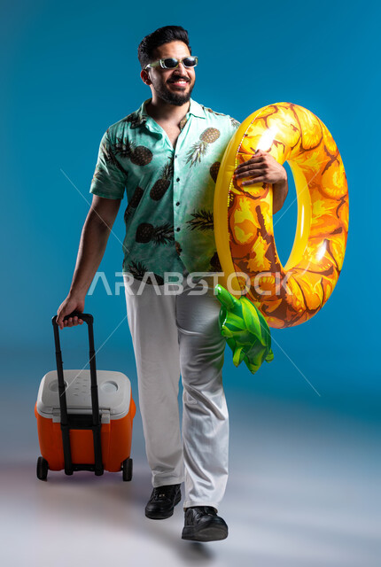 الاستمتاع بالمشروبات الباردة وقضاء وقت ممتع في العطلة الصيفية, السياحة في إحدى منتجعات السعودية, بورتريه لشاب عربي خليجي سعودي مبتسم يرتدي الزي الكاجوال والنظارة الشمسية يمسك عوامة السباحة, صورة الجسم بالكامل, خلفية زرقاء