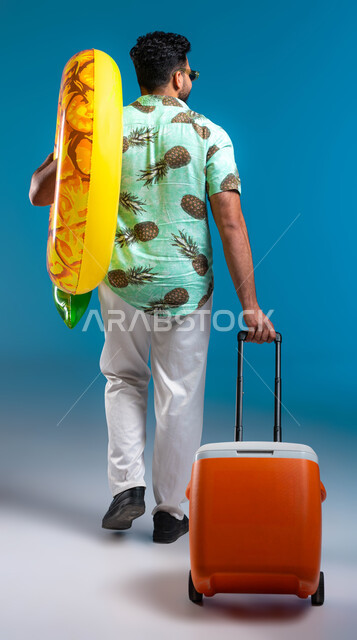 قضاء وقت ممتع في العطلة الصيفية والاستمتاع بالعصائر الباردة, السياحة في إحدى منتجعات السعودية, بورتريه من الخلف لشاب عربي خليجي سعودي مبتسم يرتدي الزي الكاجوال والنظارة الشمسية يمسك عوامة السباحة ويقوم بجر براد المشروبات, صورة الجسم بالكامل, خلفية زرقاء