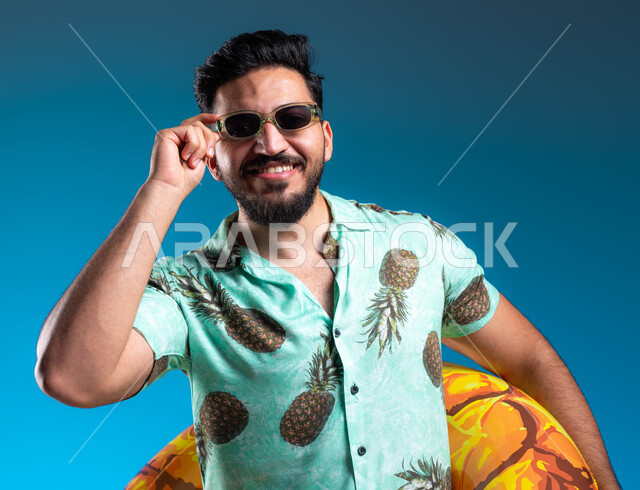 التسلية في إحدى المنتجعات الترفيهيه, بورتريه مقرب لشاب عربي خليجي سعودي مبتسم يرتدي الزي الكاجوال يمسك بيده النظارة الشمسية, قضاء أوقات ممتعه والاستمتاع بالسباحة في شواطئ السعودية بإيماءات البشاشة والسرور, خلفية زرقاء