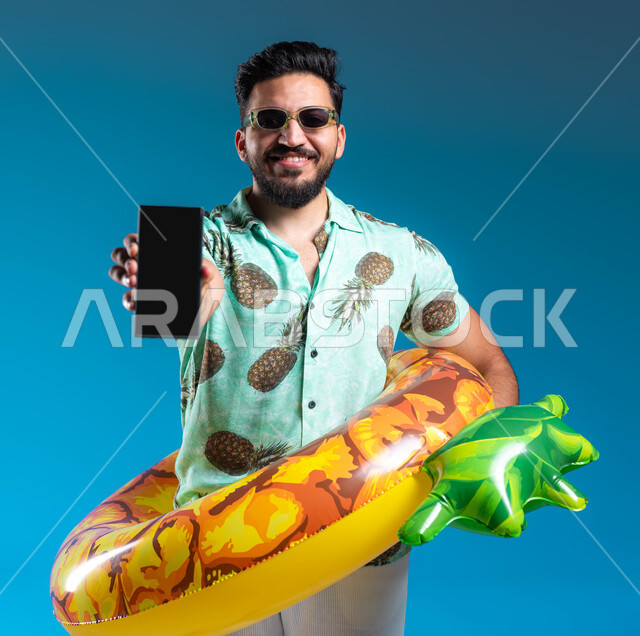 البشاشة والسرور وقضاء وقت ممتع بالعطلة الصيفية, عرض تطبيقات لخدمات الأنشطة السياحية, بورتريه لشاب عربي خليجي سعودي مبتسم يرتدي الزي الكاجوال وعوامة السباحة يحمل الهاتف المحمول بشاشة سوداء فارغة, التسليه والترفيه في إحدى المنتجعات في المملكة, خلفية زرقاء