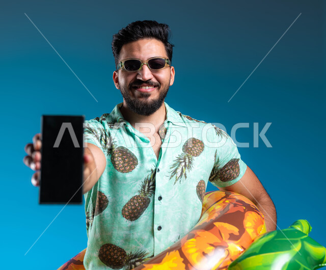 عرض تطبيقات لخدمات الأنشطة السياحية, بورتريه مقرب لشاب عربي خليجي سعودي مبتسم يرتدي الزي الكاجوال وعوامة السباحة يحمل الهاتف المحمول بشاشة سوداء فارغة, البشاشة والسرور وقضاء وقت ممتع بالعطلة الصيفية, التسليه والترفيه في إحدى المنتجعات في المملكة, خلفية زرقاء