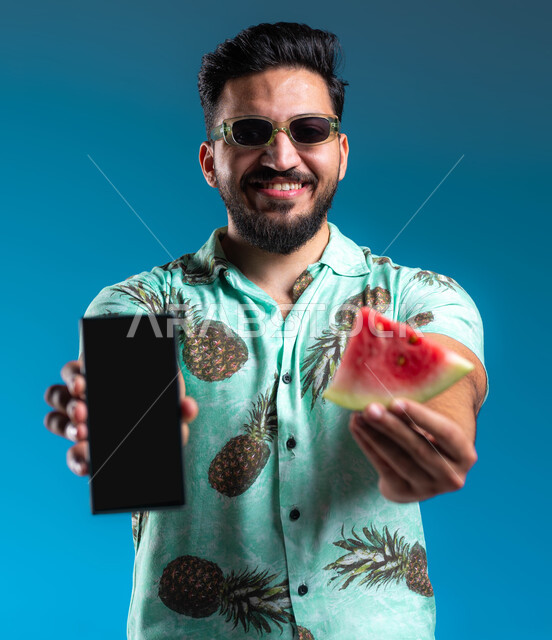 استخدام برامج وتطبيقات الهاتف المحمول, تناول الفواكه الصيفية المنعشة, بورتريه مقرب لرجل عربي خليجي سعودي مبتسم يرتدي الزي الكاجوال والنظارة الشمسية يحمل الجوال بشاشة سوداء فارغة, الاستمتاع بالإجازة السياحية في السعودية بإيماءات السرور والبشاشة, خلفية زرقاء
