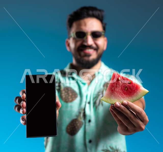 تناول الفواكه الصيفية المنعشة, استخدام برامج وتطبيقات الهاتف المحمول, بورتريه مقرب لرجل عربي خليجي سعودي مبتسم يرتدي الزي الكاجوال والنظارة الشمسية يحمل الجوال بشاشة سوداء فارغة, الاستمتاع بالإجازة السياحية في السعودية بإيماءات السرور والبشاشة, خلفية زرقاء