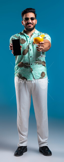 رحلة شبابية ممتعه وقضاء العطلة الصيفية مع الأصدقاء, الاستمتاع بشرب العصير الطبيعي البارد, عرض خدمات وتطبيقات السياحة والسفر, بورتريه لشاب عربي خليجي سعودي مبتسم يرتدي الزي الكاجوال والنظارة يمسك الجوال بشاشة سوداء فارغة, صورة لكامل الجسم, خلفية زرقاء