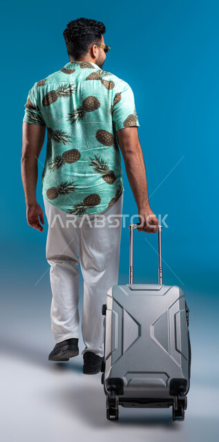 الاستمتاع في السياحة بأوقات الفراغ, الانطلاق والاستعداد لبداية عطلة صيفية سعيدة, بورتريه من الخلف لرجل عربي خليجي سعودي يرتدي الزي الكاجوال والنظارة الشمسية يقوم بجر حقيبة السفر, صورة الجسم بالطول الكامل, خلفية زرقاء
