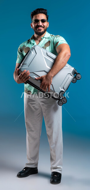 الاستعداد للرحلة السياحية ببشاشة, قضاء أوقات ممتعه خارج البلاد, بورتريه لشاب عربي خليجي سعودي مبتسم يرتدي الزي الكاجوال والنظارة يحمل حقيبة السفر بإيماءات السرور والحماس, الترفيه والتسلية والاستمتاع بالعطلة الصيفية, صورة الجسم بالطول الكامل, خلفية زرقاء