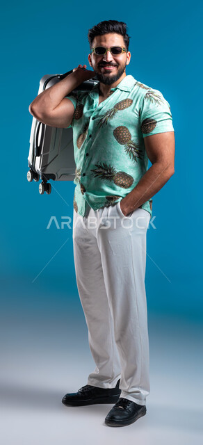 الوقوف بإيماءات الثقة والسرور والبشاشة, بورتريه لشاب عربي خليجي سعودي مبتسم يرتدي النظارة والزي الكاجوال يضع يده في جيبه ويحمل حقيبة السفر, الاستعداد للرحلة السياحية, الاستمتاع بالعطلة الصيفية وقضاء وقت ممتع مع الأصدقاء, صورة لكامل الجسم, خلفية زرقاء