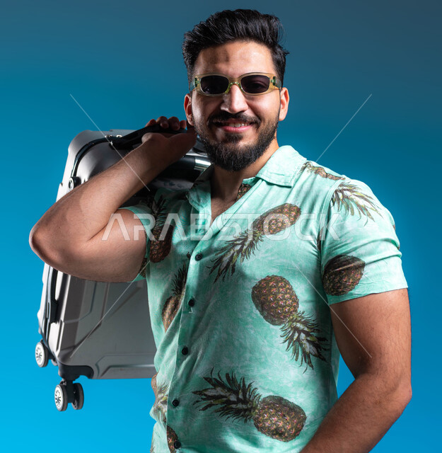 الترفيه والتسلية والاستمتاع بالعطلة الصيفية, قضاء أوقات ممتعه خارج البلاد, بورتريه مقرب لشاب عربي خليجي سعودي مبتسم يرتدي الزي الكاجوال والنظارة الشمسية يحمل حقيبة السفر بإيماءات السرور والسعادة, البشاشة والاستعداد للرحلة السياحية, خلفية زرقاء