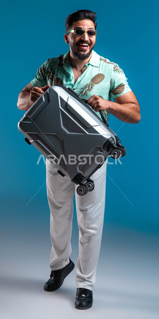 البشاشة والاستعداد للرحلة السياحية, قضاء أوقات ممتعه خارج البلاد, بورتريه لشاب عربي خليجي سعودي مبتسم يرتدي الزي الكاجوال والنظارة يحمل حقيبة السفر بإيماءات السرور والحماس, الترفيه والتسلية والاستمتاع بالعطلة الصيفية, صورة الجسم بالطول الكامل, خلفية زرقاء