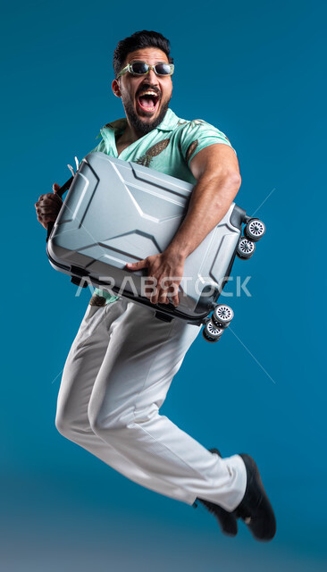 قضاء أوقات ممتعه خارج البلاد, بورتريه لشاب عربي خليجي سعودي مبتسم يرتدي الزي الكاجوال والنظارة يحمل حقيبة السفر بإيماءات القفز والحماس, البشاشة والاستعداد للرحلة السياحية, الترفيه والتسلية والاستمتاع بالعطلة الصيفية, صورة الجسم بالطول الكامل, خلفية زرقاء