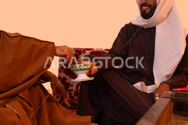 وجبة ذات قيمة غذائية عالية، الاستمتاع برحلة تخييم برية، حورارات ونقاشات بين الأصدقاء،  شاب يقوم بتقديم التمر للضيف، بورتريه مقرب لرجلان عربيان خليجيان سعوديان في كشتة شبابية، أجواء شتوية هادئة، خلفية بيج.