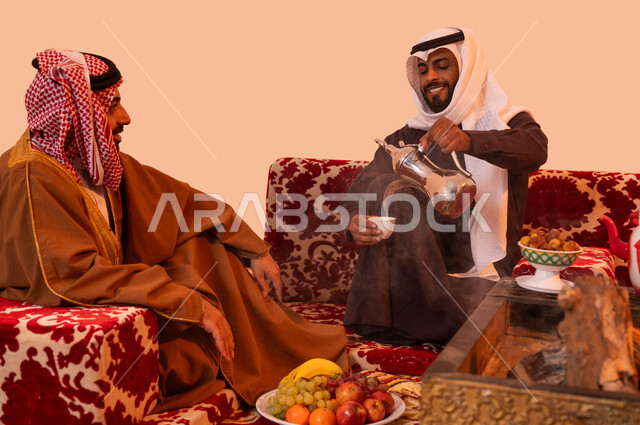 تقديم القهوة الساخنة للضيف، الاستمتاع برحلة تخييم برية، حورارات ونقاشات بين الأصدقاء،  شاب يقوم بصب القهوة للضيوف،  بورتريه لرجلان عربيان خليجيان سعوديان يرتديان الزي التقليدي يتسامران ويتبدلان الأحاديث، إيماءات البشاشة وحسن الاستقبال، خلفية بيج.
