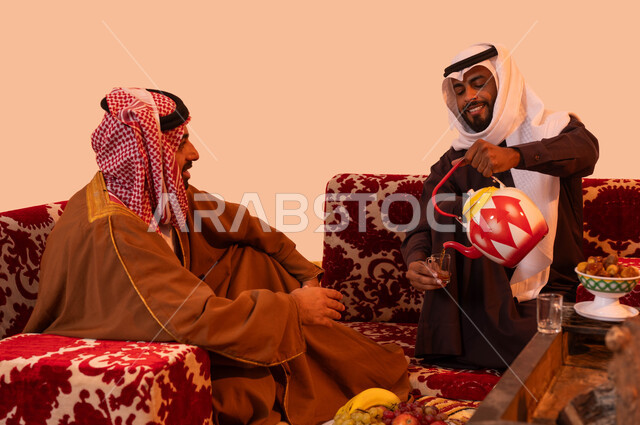 شاب يقوم بصب الشاي للضيوف، حورارات ونقاشات بين الأصدقاء، كشتة صحراوية شبابية، بورتريه مقرب لرجلان عربيان خليجيان سعوديان يرتديان الزي التقليدي يستمتعان برحلة تخييم برية، إيماءات البشاشة والفرح وحسن الاستقبال، خلفية بيج.