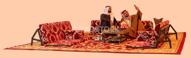 تقوية الروابط بين الأصدقاء، جلسات سعودية تقليدية في الهواء الطلق، بورتريه لصديقين عربيين خليجيين سعوديين يقومان بتوثيق اللحظات الثمينة باستخدام الهاتف المحمول، الاستمتاع بالأنشطة في الهواء الطلق أثناء الكشتة البرية، خلفية بيج