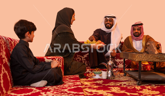 أسرة تجلس حول الموقد الطيني للتدفئة والتسامر، قضاء أوقات عائلية ممتعة، حسن الضيافه والاستقبال، إيماءات البشاشة والترحيب، بورتريه لإمرأة عربية سعودية خليجية تقوم بتقديم الفاكهة للضيوف في نزهة خارجية، الاستمتاع في الاجواء الشتوية الهادئة، خلفية بيج.