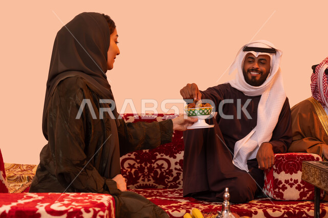 وجبة ذات قيمة غذائية عالية، نزهة برية في الصحراء، رحلات تخييم سياحية ترفيهية، مفهوم حسن الضيافه والاستقبال، بورتريه مقرب لإمرأة عربية خليجية سعودية تقوم بتقديم التمر للضيوف في جلسة خارجية، التسامر وتبادل الأحاديث في كشتة شعبية، خلفية بيج.