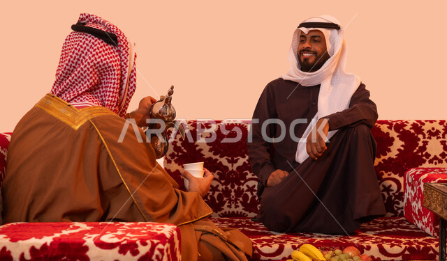 صب القهوة من الدلة النحاسية، العادات والتقاليد السعودية العريقة، إيماءات البشاشة والترحيب، مفهوم حسن الضيافه والاستقبال، التسامر وتبادل الحديث في كشتة شعبية شبابية، بورتريه مقرب لصديقان عربيان خليجيان سعوديان في جلسة بر خارجية، خلفية بيج.