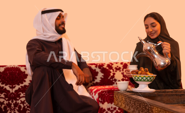 مفهوم حسن الضيافه والاستقبال، كشتة شعبية خارجية مع العائلة، عادات وتقاليد سعودية اصيلة في تقديم القهوة العربية، بورتريه مقرب لإمرأة عربية خليجية سعودية مبتسمة ترتدي العباءة تقوم بصب القهوة باستخدام الدلة النحاسية، خلفية بيج.