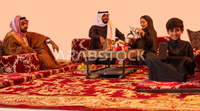 كشتة بر شعبية خارجية مع العائلة، استخدام الأجهزة والتقنيات الحديثة، مفهوم حسن الضيافه والاستقبال، بورتريه مقرب لولد عربي خليجي سعودي مبتسم يمسك الهاتف المحمول بشاشة سوداء فارغة، إمرأة ترتدي العباءة تقوم بصب القهوة للضيوف، خلفية بيج.