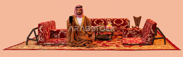 عرض برامج وتطبيقات الجوال، تقديم خدمات سياحية عبر الانترنت، مخيمات ورحلات سياحية، استخدام الأجهزة والتقنيات الحديثة، بورتريه لرجل عربي خليجي سعودي مبتسم يرتدي الشماغ والبشت التقليدي ينظر إلى الكاميرا يمسك الهاتف المحمول بشاشة سوداء فارغة، خلفية بيج.