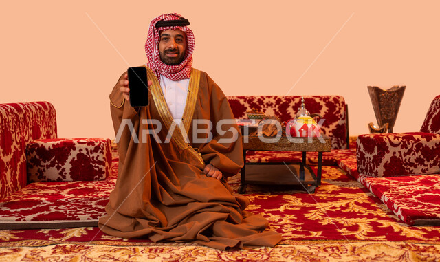 تقديم خدمات سياحية عبر الانترنت، عرض برامج وتطبيقات الجوال، مخيمات ورحلات سياحية، استخدام الأجهزة والتقنيات الحديثة، بورتريه لرجل عربي خليجي سعودي مبتسم يرتدي الشماغ والبشت التقليدي ينظر إلى الكاميرا يمسك الهاتف المحمول بشاشة سوداء فارغة، خلفية بيج.