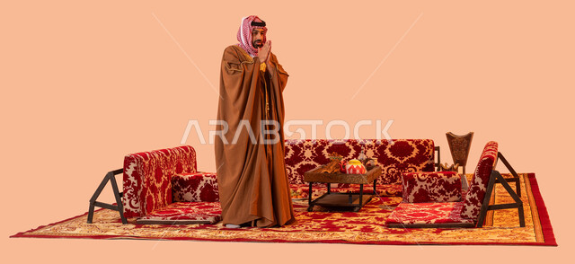 الأجواء الشتوية الباردة في أماكن التخييم، الشعور بالصقيع والبرد الشديد، بورتريه لرجل عربي خليجي سعودي يرتدي البشت والشماغ التقليدي ويضع يديه على كتفه بشكل متقاطع، صورة الجسم بالطول الكامل، خلفية بيج .