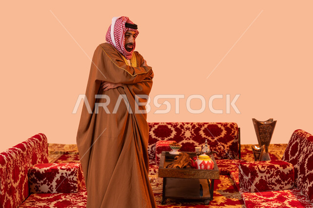 الشعور بالصقيع والبرد الشديد، بورتريه لرجل عربي خليجي سعودي يرتدي البشت والشماغ التقليدي ويضع يديه على كتفه بشكل متقاطع، الشعور بالصقيع، الأجواء الشتوية الباردة في أماكن التخييم، صورة الجسم بالطول الكامل، خلفية بيج .