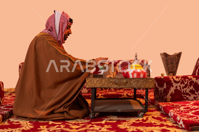 أسلوب الحياة البدوية القديمة, جلسة بر شبابية شعبية، إيماءات التدفئة على الموقد, مخيمات وأجواء شتوية، بورتريه مقرب لرجل عربي خليجي سعودي يرتدي البشت والشماغ التقليدي يجلس أمام شعلة من النار ، الأجواء الهادئة في أماكن التخييم، خلفية بيج.