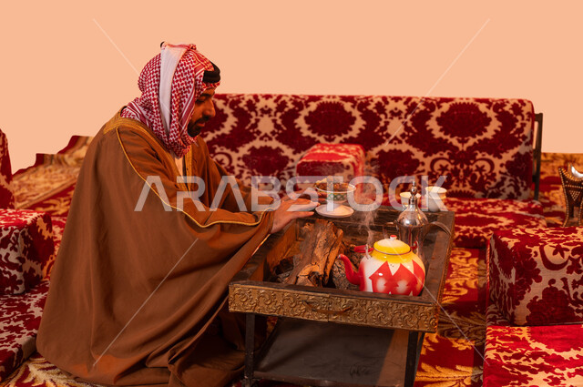 الأجواء الهادئة في أماكن التخييم، جلسة بر شبابية شعبية، أسلوب الحياة القديمة، إيماءات التدفئة على الموقد, مخيمات وأجواء شتوية، بورتريه مقرب لرجل عربي خليجي سعودي يرتدي البشت والشماغ التقليدي يجلس أمام شعلة من النار ، خلفية بيج.