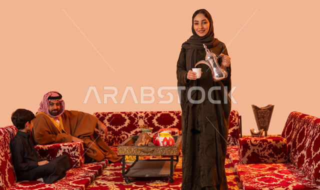 مفهوم حسن الضيافه والكرم، جلسات شعبية خارجية مع العائلة، عادات وتقاليد سعودية اصيلة في تقديم القهوة العربية، بورتريه لإمرأة عربية خليجية سعودية مبتسمة ترتدي العباءة تقوم بصب القهوة باستخدام الدلة النحاسية، صورة الجسم بالطول الكامل، خلفية بيج.