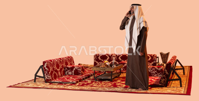 استخدام التقنيات والوسائل الحديثة والمطورة للتواصل مع الأهل والأصدقاء، رحلة تخييم في الصحراء السعودية، بورتريه لرجل عربي خليجي سعودي كبير بالسن يرتدي الزي التقليدي يجري مكالمة هاتفية عبر الجوال، صورة الجسم بالكامل، خلفية بيج.