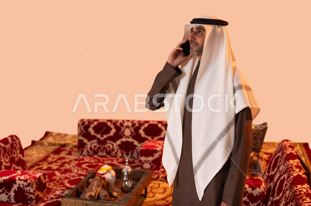 Communicating with family and friends via mobile phone, popular Saudi-style sessions, using modern and developed technologies and means, a close-up portrait of an elderly Saudi Gulf Arab man wearing traditional clothing making a mobile phone call, beige background.