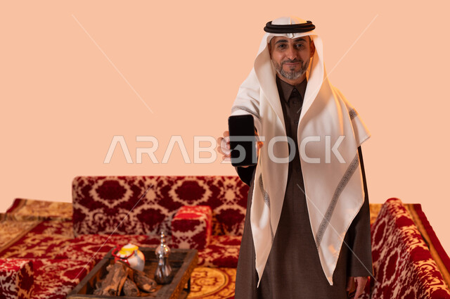 جلسات شعبية على الطراز السعودي، استخدام التقنيات والوسائل الحديثة والمطورة، بورتريه مقرب لرجل عربي خليجي سعودي كبير بالسن مبتسم يرتدي الزي التقليدي يحمل بيده الهاتف الجوال بشاشة سوداء فارغة، خلفية بيج.