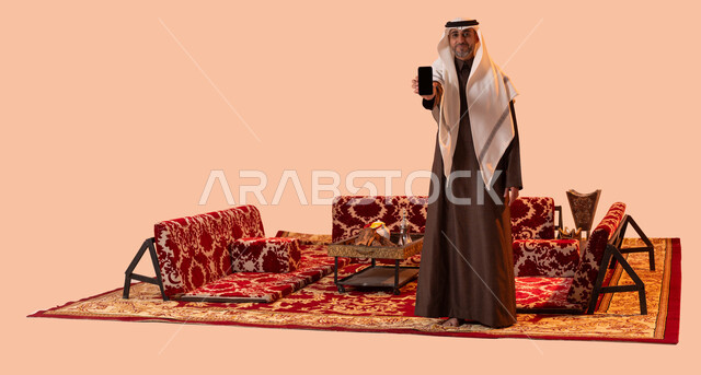 استخدام التقنيات والوسائل الحديثة والمطورة، عرض شاشه سوداء فارغة، جلسات عربية شعبية خارجية، بورتريه لرجل عربي خليجي سعودي كبير بالسن مبتسم يرتدي الزي التقليدي يحمل بيده الهاتف الجوال، صورة لكامل الجسم، خلفية بيج.